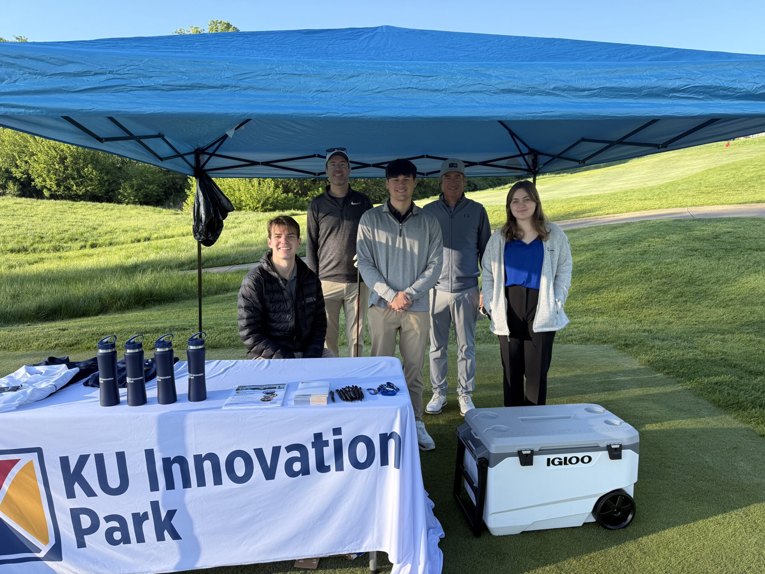 Five people pose together under a blue canopy tent on a golf course, standing behind a KU Innovation Park table. The table is covered with branded materials including navy water bottles, pens, and brochures. A large Igloo cooler sits nearby on the grass. The group is casually dressed for an outdoor event, with a sunny, green landscape in the background.